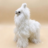 A fluffy white alpaca plush toy with long shaggy fur, small black eyes, and a small white nose, standing against a plain beige studio background.