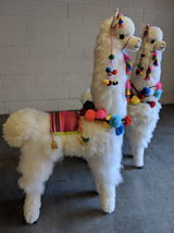 A handcrafted white llama made from alpaca fur, adorned with colorful pompoms and decorative trims, standing on a flat surface.