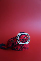 A red and black hand woven belt with floral embroidery and a metallic buckle.