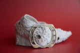 A hand woven wool belt with white embroidery and a gold-colored buckle on a red background.